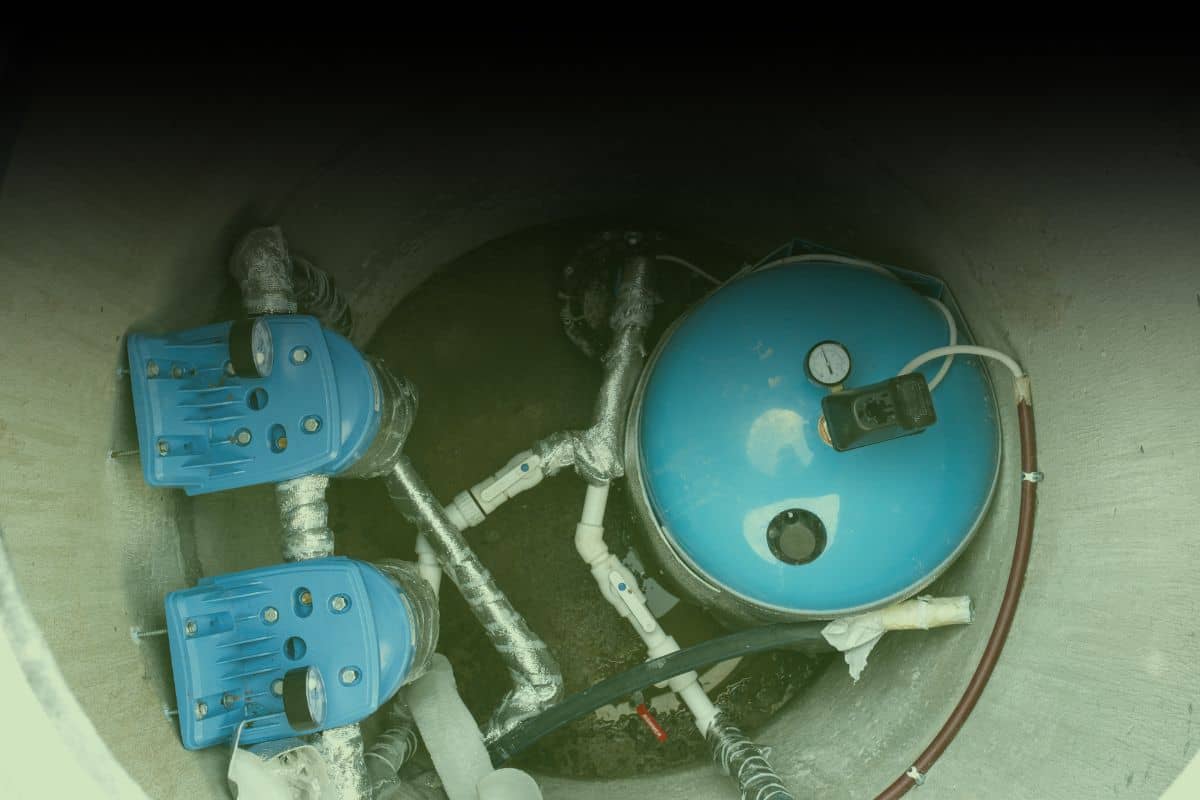 Inside a concrete well pit showing a pressure tank, dual water pumps, and plumbing lines during a professional water well inspection by Inspection Gator.