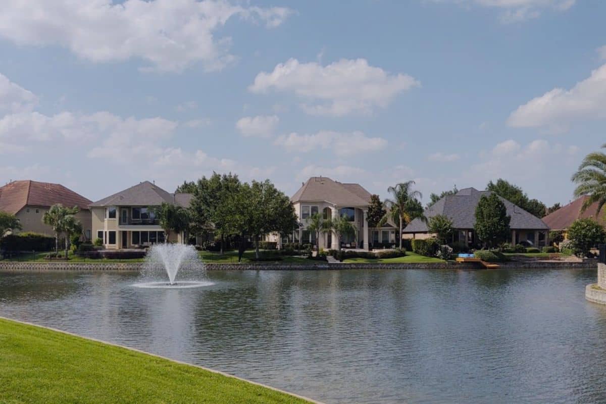 Waterfront homes along a Texas lake with a fountain, mature trees, and green lawns under a blue sky