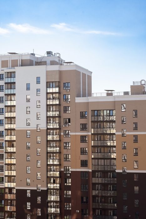 Modern multi-story residential building with a beige, white, and brown facade featuring glass balconies under a blue sky.