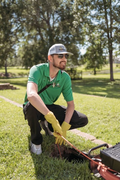 Home inspector wearing rubber gloves feeding an orange sewer camera cable into an underground pipe access on a lawn.